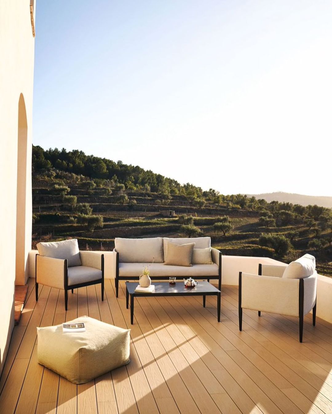 A white grey outdoor sofa set with 2 armchairs near a black coffee table and a pouf on a veranda near a vineyard.